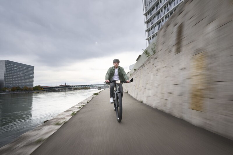 A man wearing a helmet rides an e-bike in front of a stone wall while the sky is overcast.